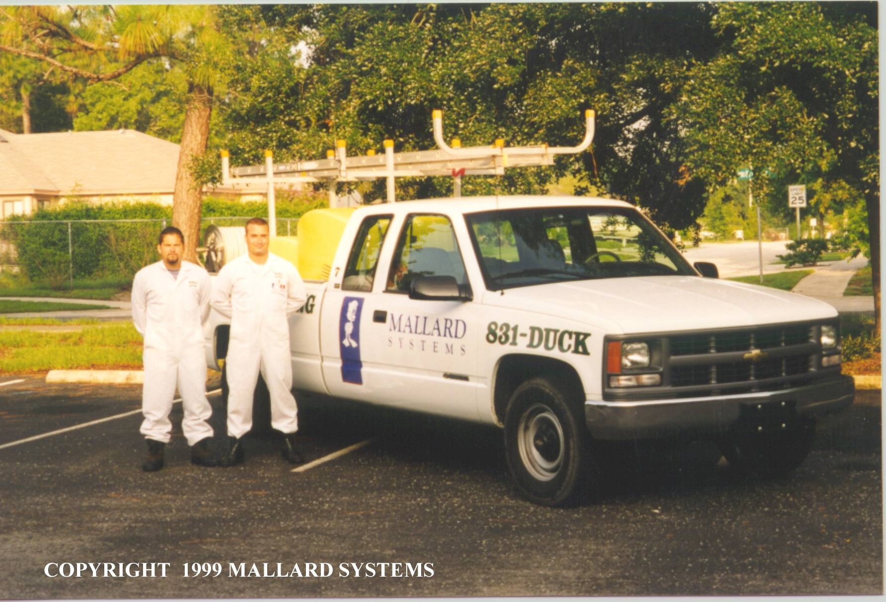 Two technicians in white coveralls stand beside a white Mallard Systems service truck with ladder rack in a parking lot