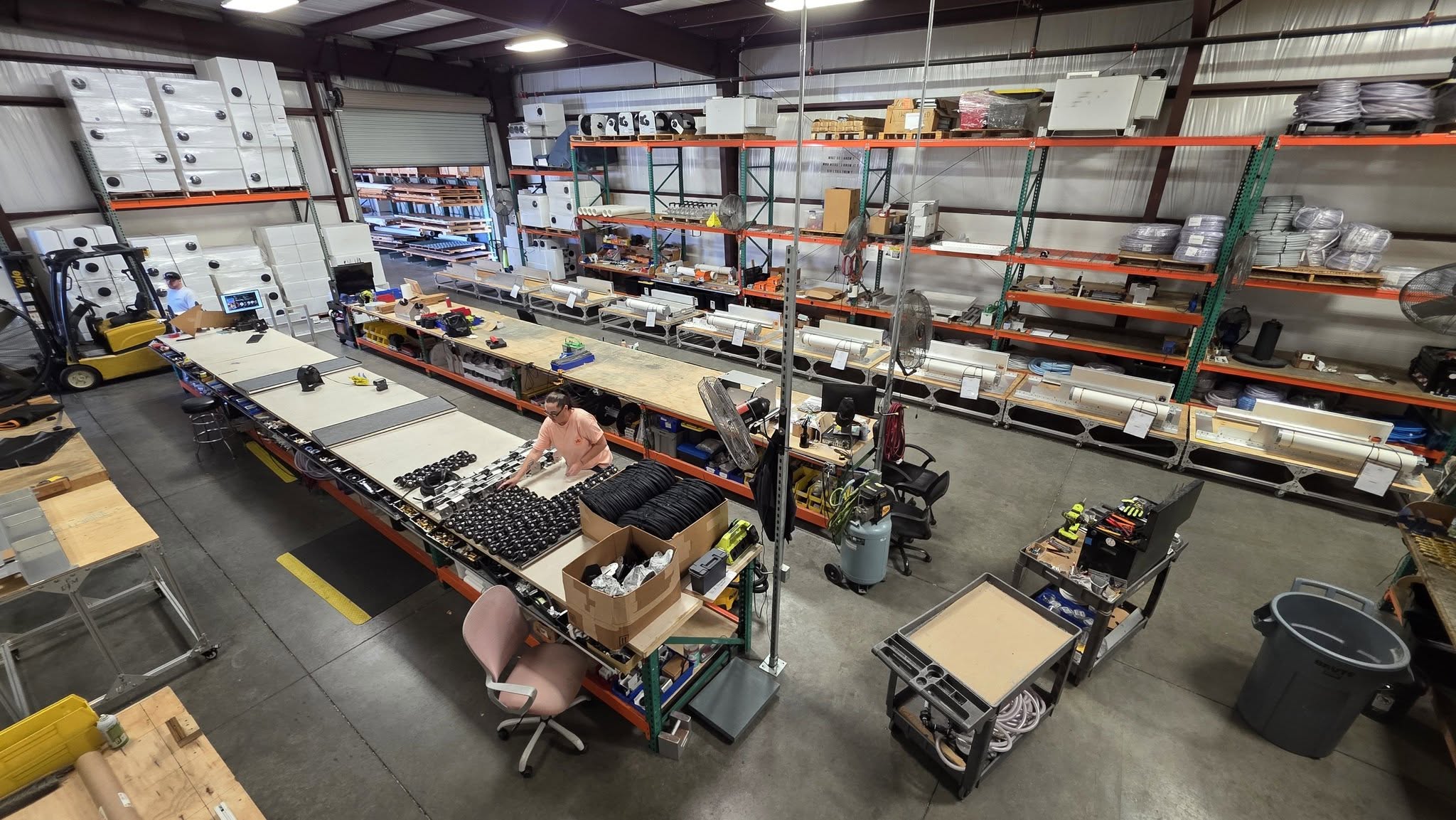 Industrial workshop with a long workbench, shelves filled with parts, and two workers organizing components