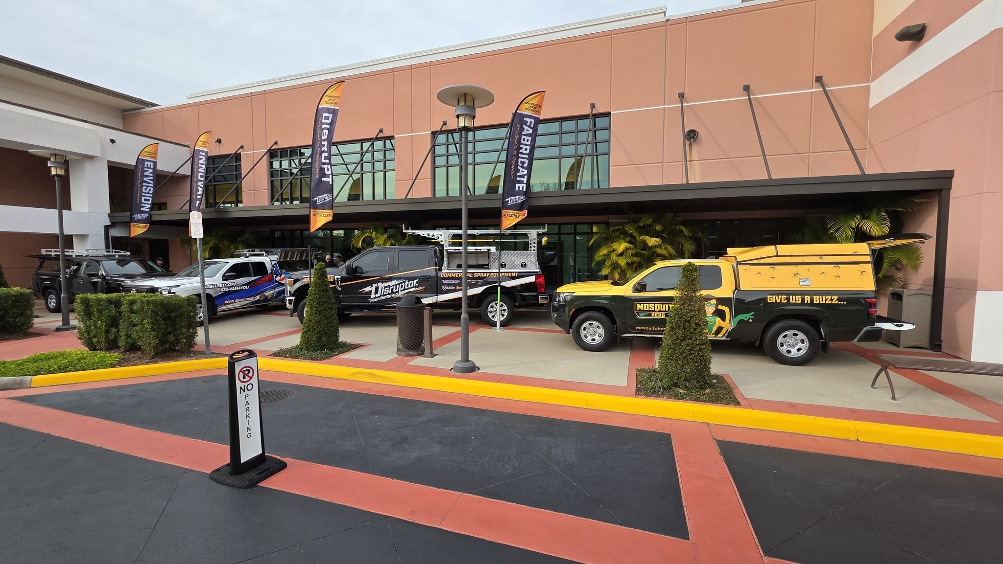 615436840_1442646801204678_9189803445426649387_n Promotional vehicles parked outside a peach-colored commercial building with tall orange-and-black banners along the storefront façade.