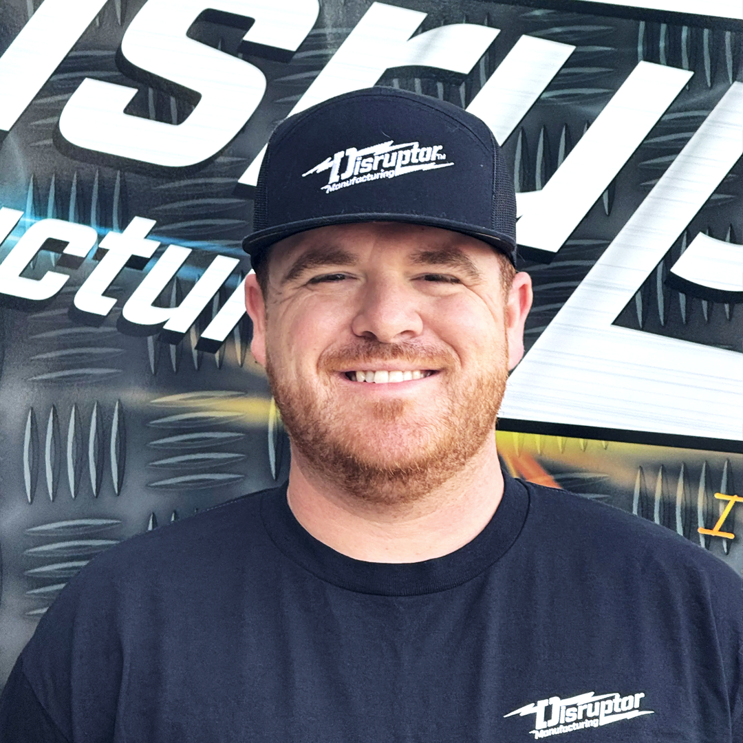 Smiling man wearing a black cap and T‑shirt with a Disruptor Manufacturing logo, standing in front of a metal-patterned backdrop.