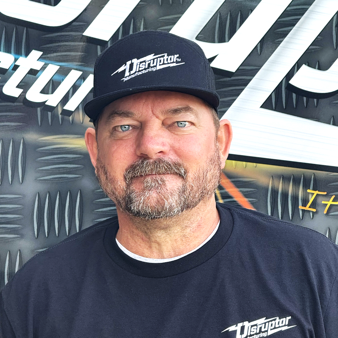 Portrait of a man with blue eyes and a goatee wearing a black cap with the Disruptor Manufacturing logo, standing in front of a diamond-plate background.