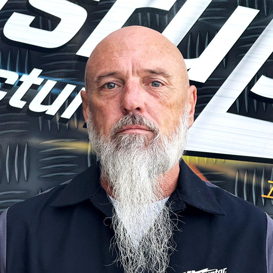 Bald man with a long white beard wearing a dark vest, looking straight at the camera with a neutral expression against a graphic backdrop.