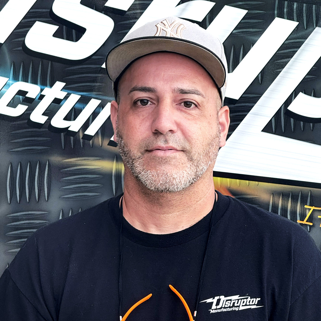 Man in a black Disruptor Manufacturing t-shirt and gray cap, standing in front of a diamond-plate backdrop with the word 'Disruptor' visible.
