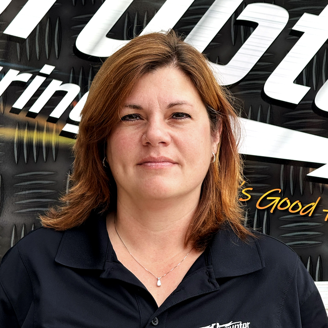 Head-and-shoulders portrait of a woman with light brown hair wearing a black polo shirt, standing in front of a black metal backdrop with white lettering.