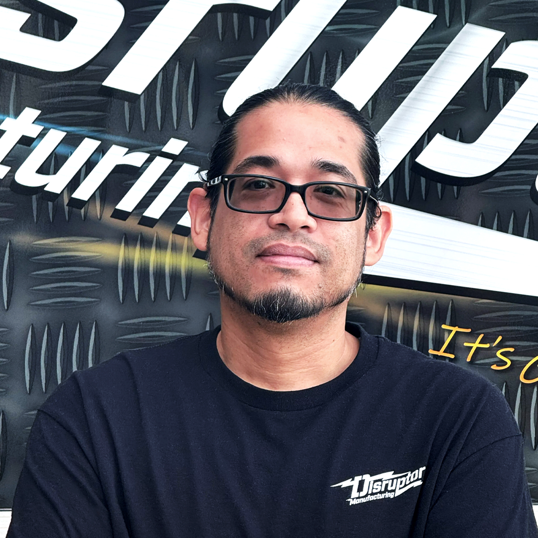 Man in a black Disruptor Manufacturing t-shirt and gray cap, standing in front of a diamond-plate backdrop with the word 'Disruptor' visible.