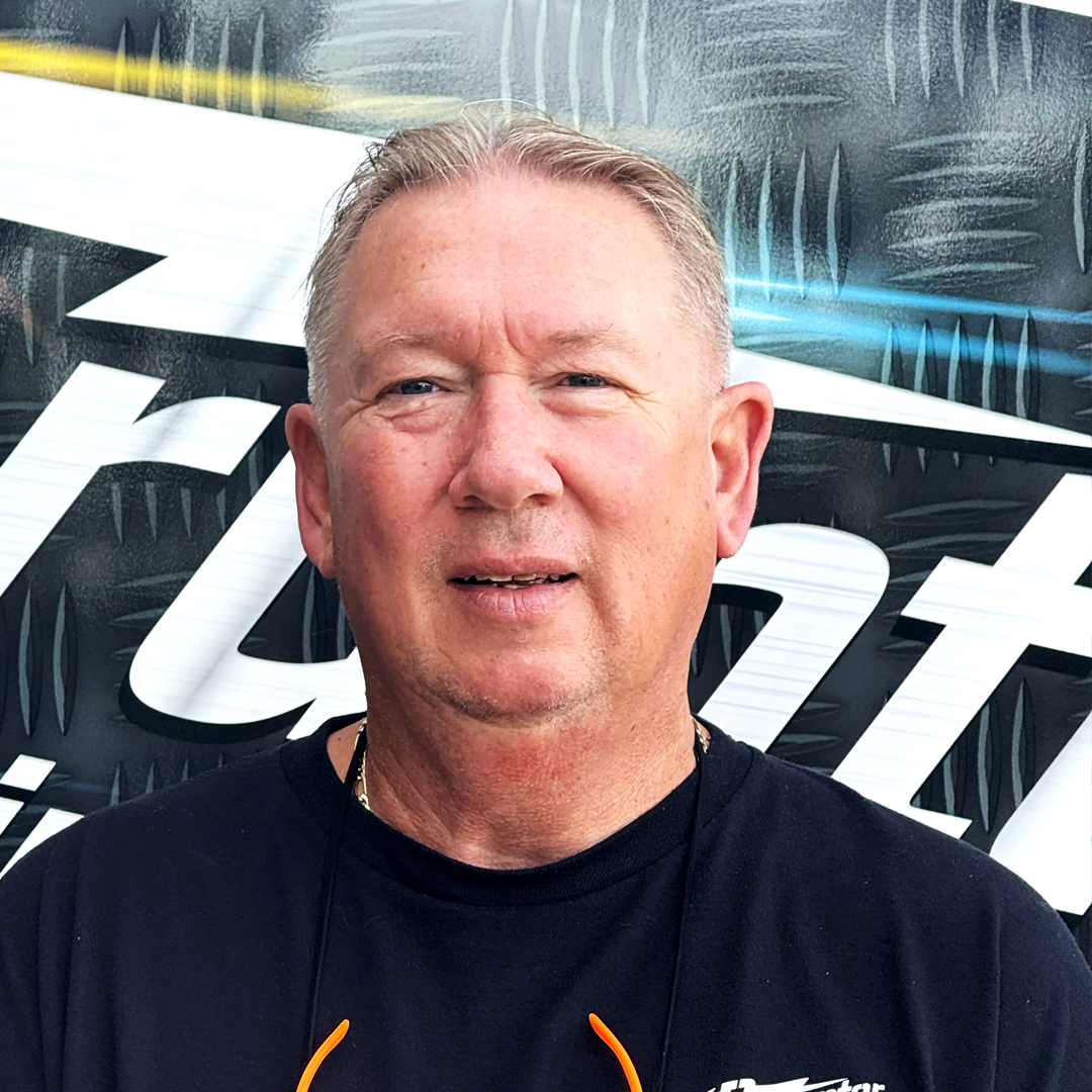 Portrait of a middle-aged man with short gray hair, wearing a black T-shirt and orange sunglasses hanging from his shirt, standing in front of a dark, graphic vehicle surface with metallic patterns.