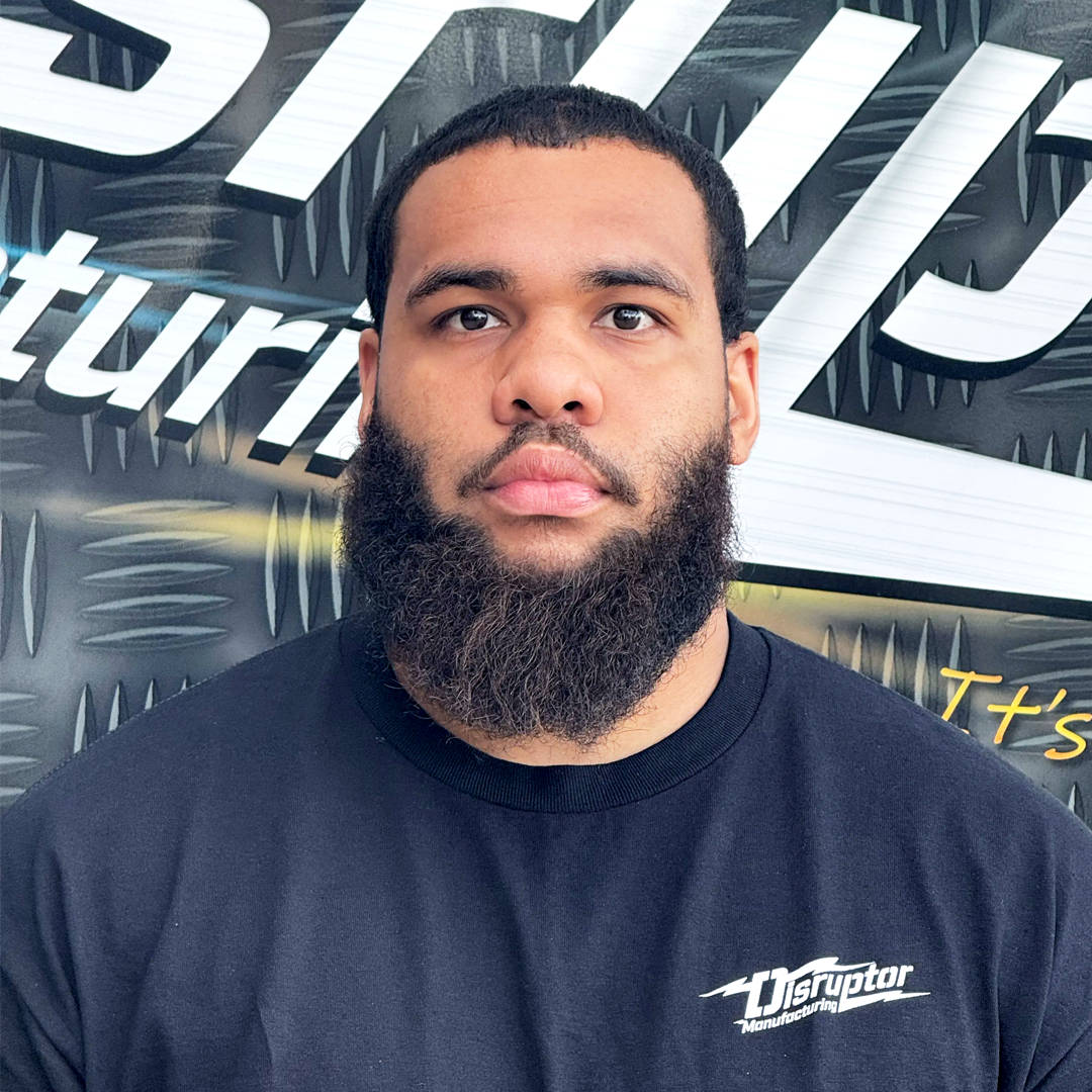 Portrait of a man with glasses and a full beard wearing a black shirt with a Disruptor Manufacturing logo, standing in front of a textured backdrop.